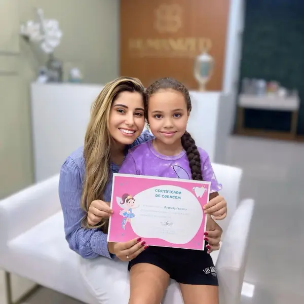 Dentista e paciente mirim segurando juntas o certificado de coragem na recepcao da clinica